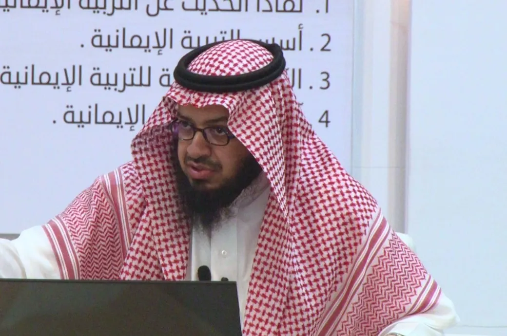 فايز الزهراني بشماغٍ أحمر ونظارة يحاضر ويشير بيده، وخلفه شاشة عرضٍ عن التربية الإيمانية