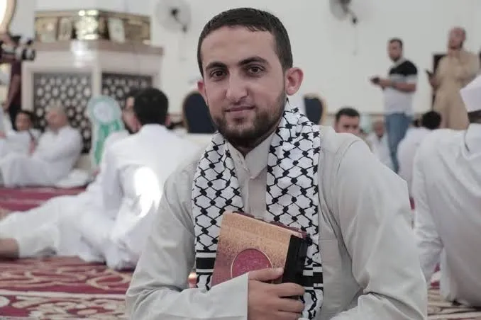 محمد زكي حمد في مسجدٍ مفروش، يضمّ مصحفًا إلى صدره وعلى كتفيه كوفية، وخلفه مصلّون