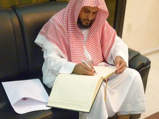 الشيخ عبد العزيز الطريفي جالسًا على أريكة جلدية، يكتب بقلمه في سجلٍّ كبير مفتوح على ركبتيه