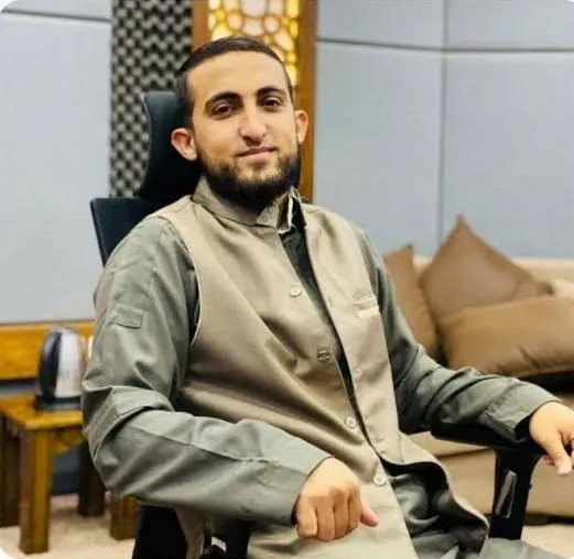 محمد زكي حمد جالسًا على كرسيّ مكتب بثوبٍ وصديريّة، مبتسمًا