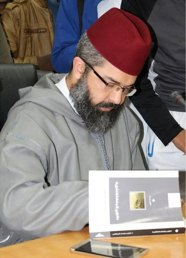 البشير عصام المراكشي بطربوشٍ أحمر وجلبابٍ رمادي يوقّع كتابًا، والناس وقوفٌ حوله