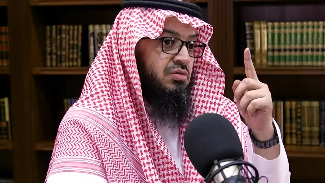 فايز الزهراني بشماغٍ أحمر ونظارة يرفع سبّابته متحدّثًا أمام مكبّر صوت، وخلفه رفوف كتب