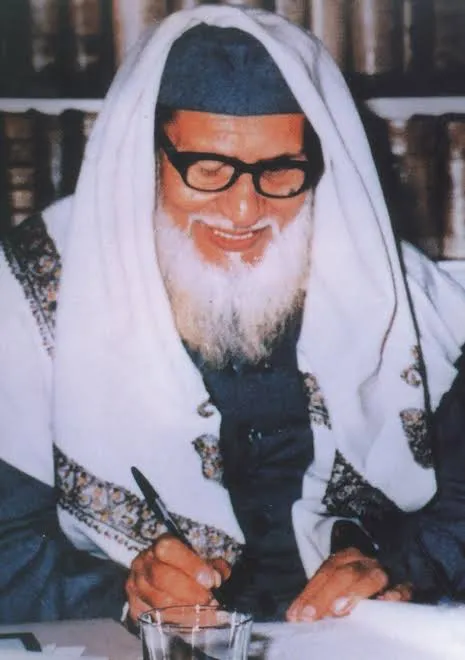 أبو الحسن الندوي يكتب مبتسمًا أمام رفوف الكتب