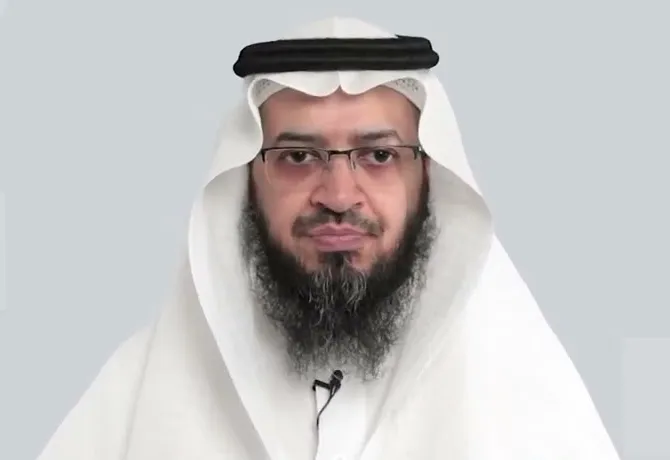 فايز الزهراني بغترةٍ بيضاء ونظارة، على صدره مكبّر صوتٍ صغير، أمام خلفيةٍ رمادية
