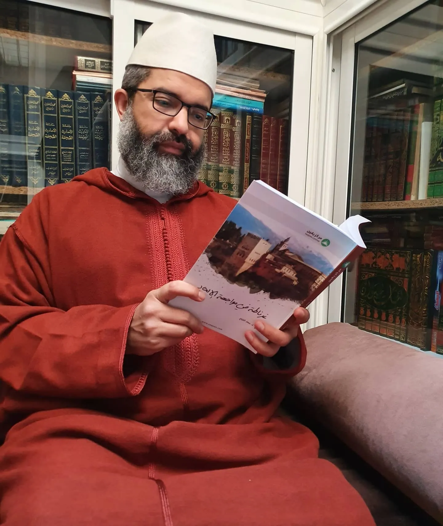 البشير عصام المراكشي بطاقيةٍ بيضاء وجلبابٍ أحمر يقرأ كتابًا، وخلفه خزائن كتب