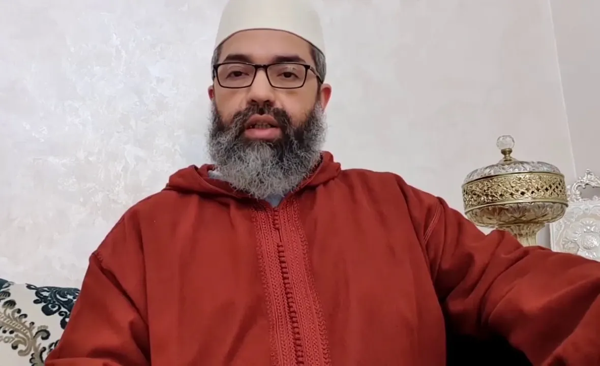 البشير عصام المراكشي بطاقيةٍ بيضاء وجلبابٍ أحمر يتحدّث إلى الكاميرا في غرفةٍ بيضاء