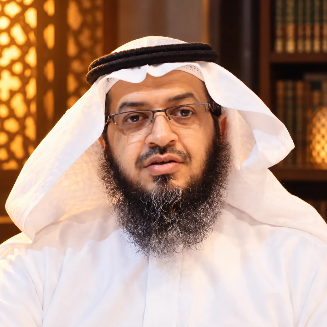 صورة فايز بن سعيد الزهراني بغترةٍ بيضاء ونظارة، وخلفه مشربيّة مضاءة ورفوف كتب